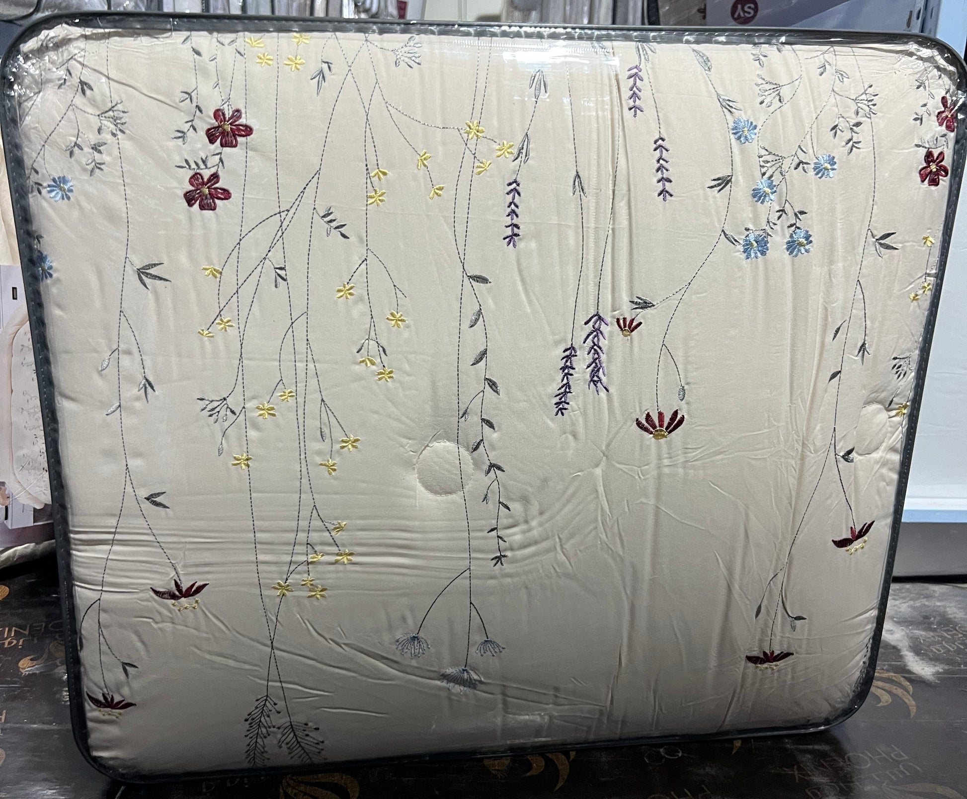 A square quilted mattress topper or mattress panel with a cream-colored floral pattern, wrapped in protective plastic packaging and standing on a dark floor.